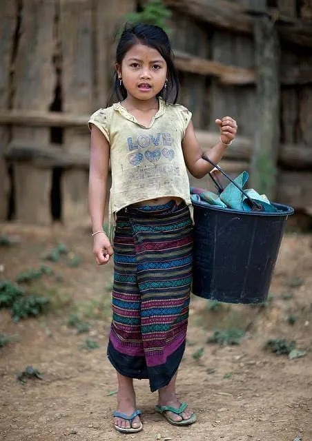 A child standing outside a wooden house holding a bucket