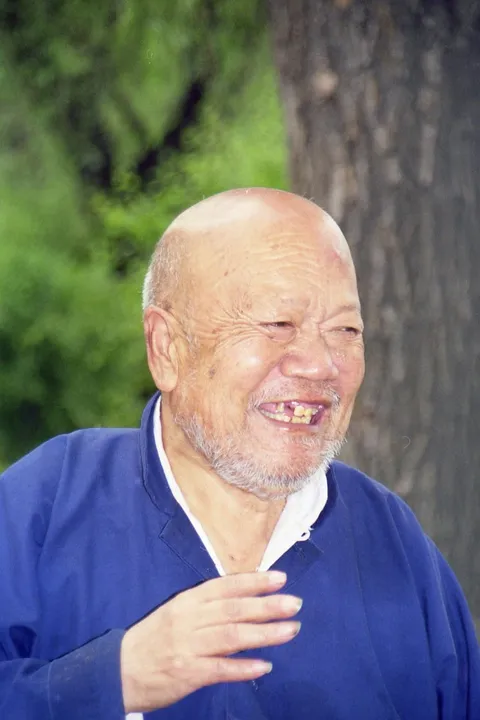 An older man in a blue jacket smiling and holding a cigarette