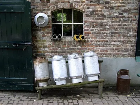 Metal milk churns on a wooden bench outside a brick farmhouse