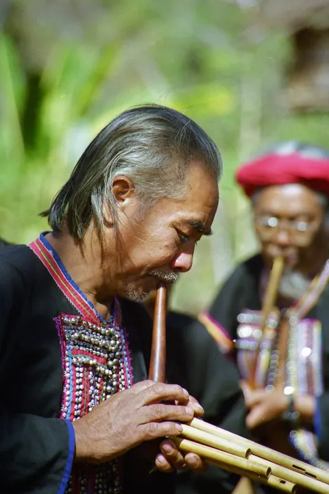 A grey-haired man in an embroidered black jacket playing a bamboo wind instrument