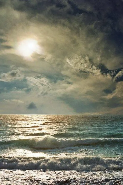 Sun shining through heavy clouds over waves breaking on a shore