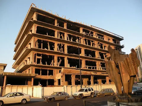 Gutted multi-storey concrete building beside a bronze flame-shaped sculpture, cars parked on the street below