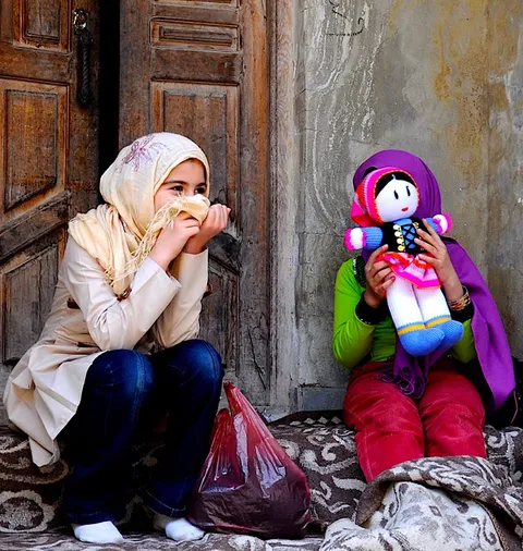 A woman and child sitting outside a wooden door, the child holding a doll