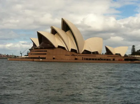 A building with white sail-shaped roofs across the water under cloudy skies