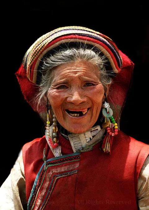 A smiling elderly woman in a striped headdress, beaded earrings and red vest