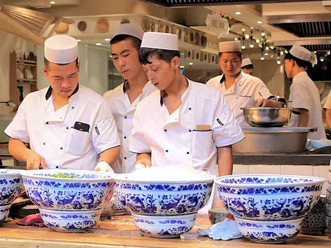 Cooks in white uniforms working behind bowls of food at a counter