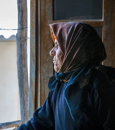 An older woman in a headscarf looking out of a window