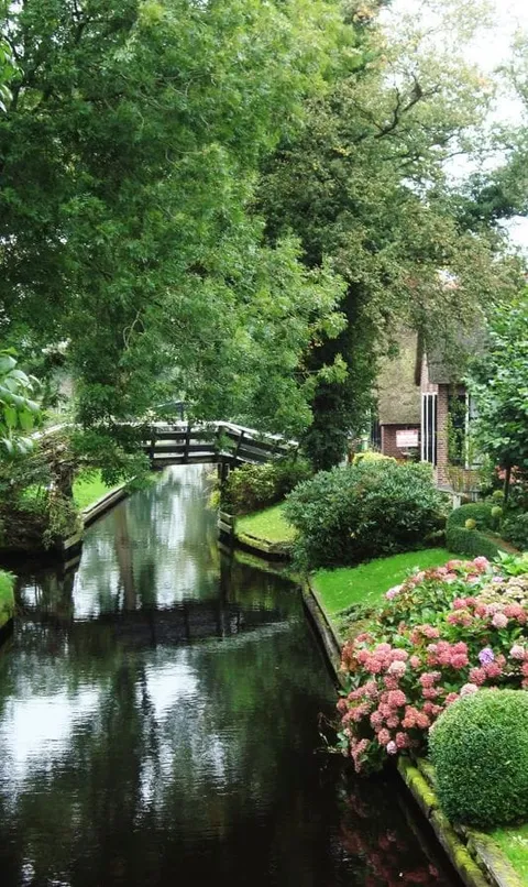 A narrow canal beneath a small footbridge with flowers along the bank