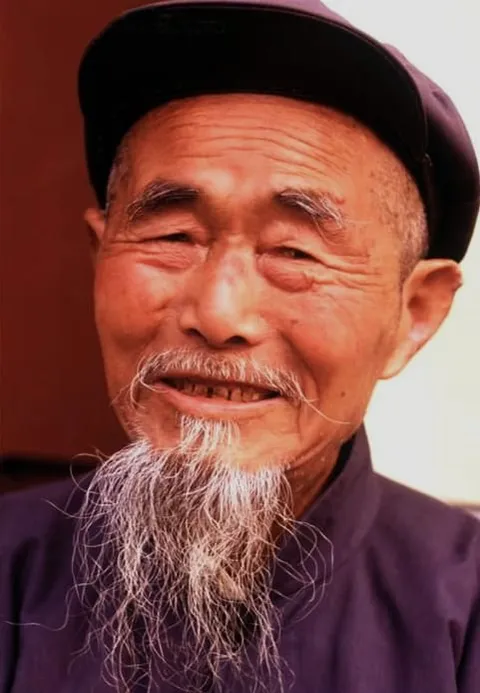 A smiling elderly man with a long white beard wearing a dark cap