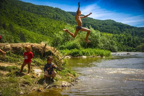 Children playing beside a river, one jumping from the grassy bank