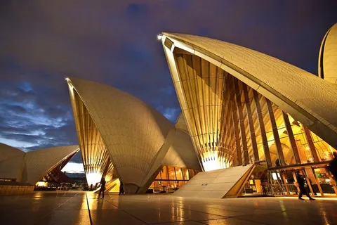 Illuminated sail-shaped roofs of a building at dusk with people walking