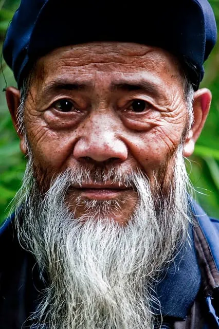 An elderly man with a long white beard and dark cap in front of foliage