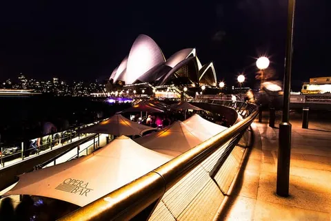 A waterfront promenade with white umbrellas and a lit sail-roofed building at night