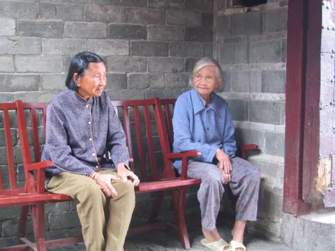 Two older men sitting together on a wooden bench against a brick wall