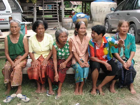 Six older women sitting barefoot on a wooden bench on grass