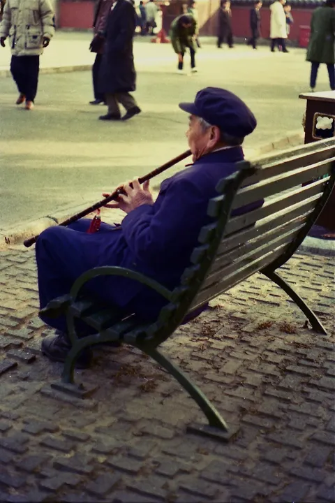 An elderly man in a blue cap playing a flute on a park bench