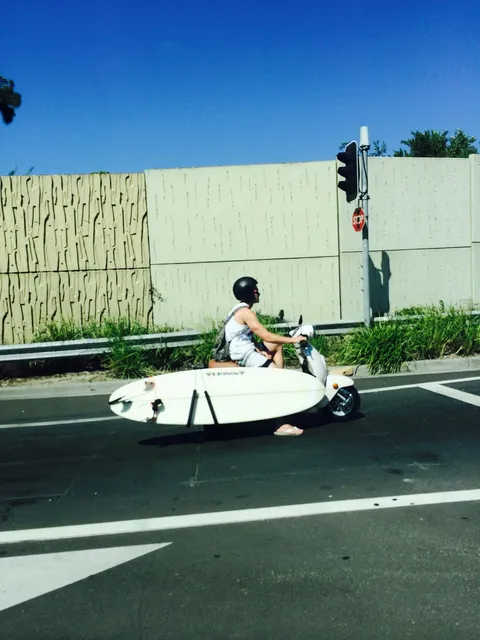 A man riding a scooter with a surfboard strapped to its side