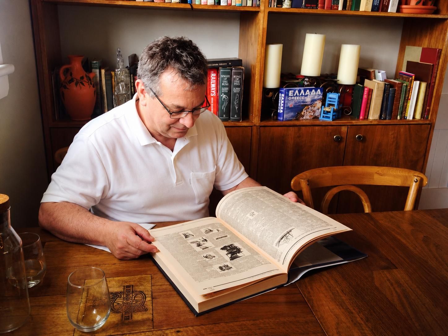 Stephanus Peters, wearing glasses and a white polo shirt, reading a very large open book at a wooden table, with a bookshelf behind him